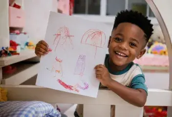 A child smiling while holding up a drawing in a classroom